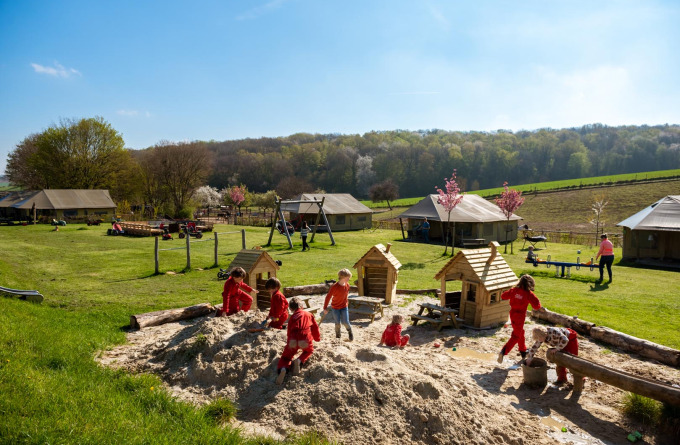 Kinderen spelen in een grote zandbak op FarmCamps De Verloren Kost met grasvelden en tenten op de achtergrond.