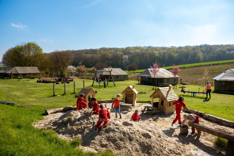 Niños juegan en una gran caja de arena en FarmCamps De Verloren Kost, con prados y tiendas al fondo.
