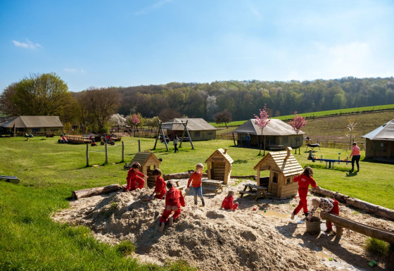 Niños juegan en una gran caja de arena en FarmCamps De Verloren Kost, con prados y tiendas al fondo.