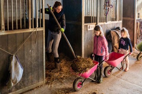 Eine Frau und zwei Kinder schaufeln Heu in Schubkarren in einem Stall im FarmCamps De Verloren Kost.