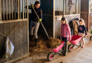 En kvinde og to børn arbejder sammen med hø og trillebøre i en stald på FarmCamps De Verloren Kost.