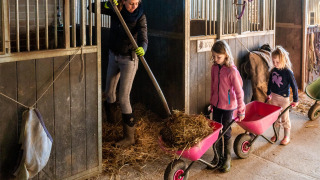 Una mujer y dos niñas trabajan juntas en un establo con carretillas y paja en FarmCamps De Verloren Kost.