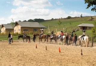 Bambini a lezione di equitazione al FarmCamps De Verloren Kost in Limburgo, Paesi Bassi, con tende dietro.