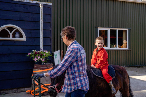 Un niño con traje rojo monta un pony guiado por un adulto en FarmCamps Five Star en Zelanda, Países Bajos.