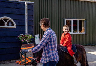 Een jongen in rode overall rijdt op een pony, begeleid door een volwassene op FarmCamps Five Star in Zeeland.