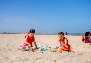 Twee meisjes in oranje jurkjes spelen met emmers en schepjes op het strand bij FarmCamps Five Star in Zeeland.