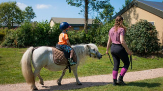 Un niño montando un pony pequeño, guiado por un adulto en FarmCamps Fleur Stables en Holanda Septentrional.