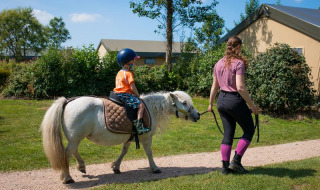 Un niño montando un pony pequeño, guiado por un adulto en FarmCamps Fleur Stables en Holanda Septentrional.