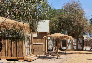 Udendørsområde i TAIGA Almeria Playa med træhytter, stråtage og stole under solrige forhold i Andalusien.