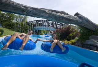 Two people relax on blue inflatable tubes in a lazy river at Camping Terme Catez, Slovenia, below a log bridge.