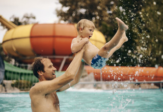 Un uomo gioca con un bambino in piscina al Camping Terme Catez, Slovenia, con scivolo d’acqua sullo sfondo.