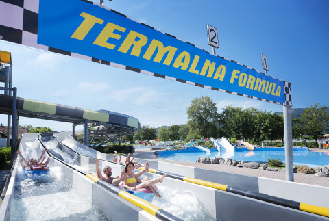 People riding water slides below the 'Termalna Formula' sign at Camping Terme Čatež in Slovenia on a sunny day.