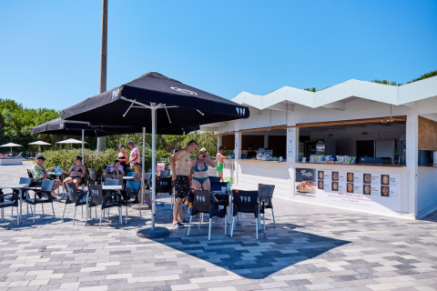 Outdoor snack bar with umbrellas and people relaxing at Camping Village Baia Blu La Tortuga in Sardinia, Italy.