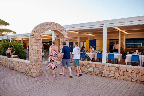 Huéspedes cruzan un arco de piedra hacia el restaurante en Camping Village Baia Blu La Tortuga, Cerdeña, Italia.