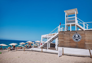 Beach scene at Camping Village Baia Blu La Tortuga in Sardinia with lifeguard tower and sun umbrellas.