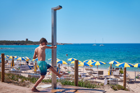 Niño usando la ducha junto a la playa en Camping Village Baia Blu La Tortuga, Cerdeña, Italia, con sombrillas.