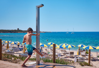Dreng tager brusebad ved stranden i Camping Village Baia Blu La Tortuga, Sardinien, Italien, med farverige parasoller.