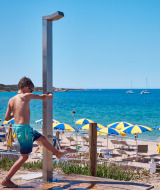 Niño usando la ducha junto a la playa en Camping Village Baia Blu La Tortuga, Cerdeña, Italia, con sombrillas.