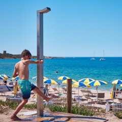 Niño usando la ducha junto a la playa en Camping Village Baia Blu La Tortuga, Cerdeña, Italia, con sombrillas.