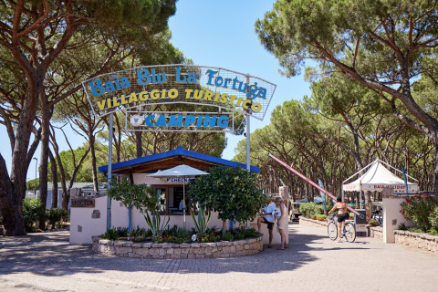 Entrada al Camping Village Baia Blu La Tortuga en Cerdeña, Italia, rodeada de pinos y visitantes turísticos.