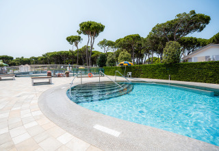 Udendørs swimmingpool med trappe, bænke og grønne træer i Camping Village Cavallino, Veneto, Italien.