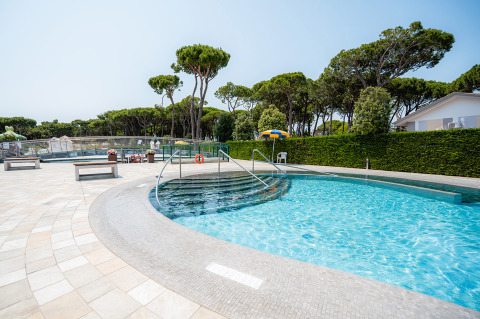 Udendørs swimmingpool med trappe, bænke og grønne træer i Camping Village Cavallino, Veneto, Italien.