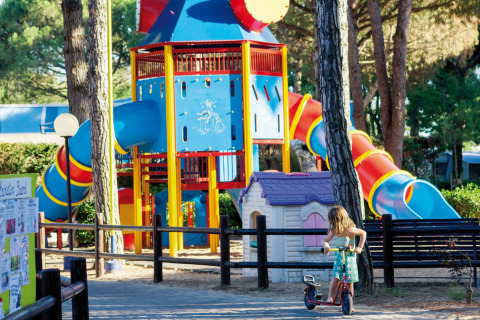 Area giochi colorata a Camping Village Cavallino, Veneto, Italia, con scivoli e bambina su monopattino.