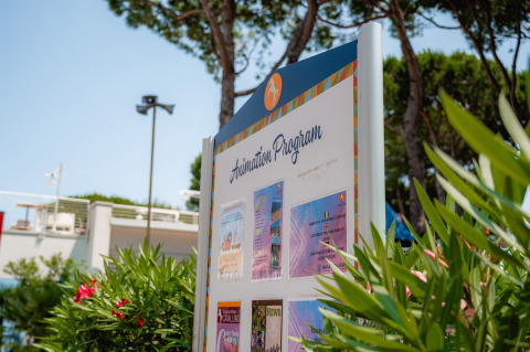 Signboard showing animation program at Camping Village Cavallino, Veneto, Italy, among lush greenery.