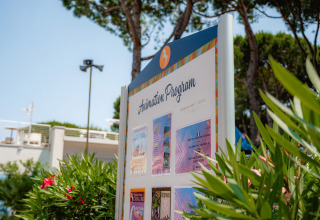 Signboard showing animation program at Camping Village Cavallino, Veneto, Italy, among lush greenery.