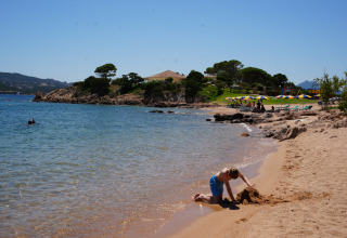 Dreng bygger sandslot på stranden ved Centro Vacanze Isuledda, omgivet af krystalklart vand og parasoller.