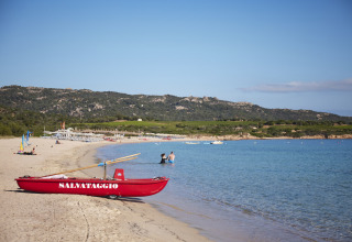 Solrig strand ved Centro Vacanze Isuledda i Sardinien, Italien med redningsbåd og bjergrigt landskab.