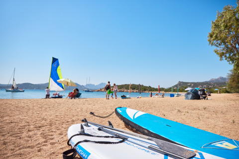 Strandscene ved Centro Vacanze Isuledda i Sardinien, Italien, med surfbrætter, badegæster og sejlskibe.