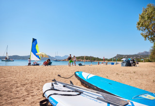 Playa en Centro Vacanze Isuledda, Cerdeña, Italia, con tablas de surf, bañistas y barcos de vela.