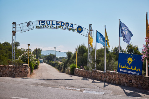Entrada al Centro Vacanze Isuledda, parque vacacional en Cerdeña, Italia, con banderas y vegetación.
