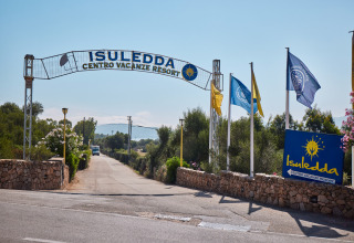 Indgangen til Centro Vacanze Isuledda feriepark i Sardinien, Italien med flag og vej omringet af grønne træer.