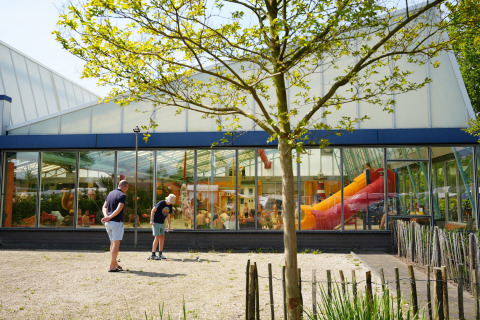 To personer spiller petanque udenfor en bygning med store vinduer og orange rutsjebane i Holiday Park Ackersate.