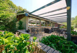 Patio esterno coperto con tavoli e sedie presso Holiday Resort de Bronzen Emmer, circondato dal verde a Drenthe.