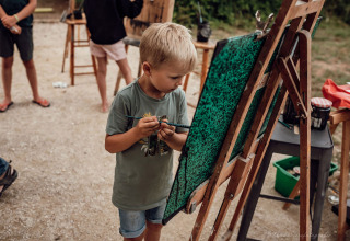 Ein kleiner Junge malt konzentriert an einer Staffelei im Freien bei Medrose Camping in Frankreich.