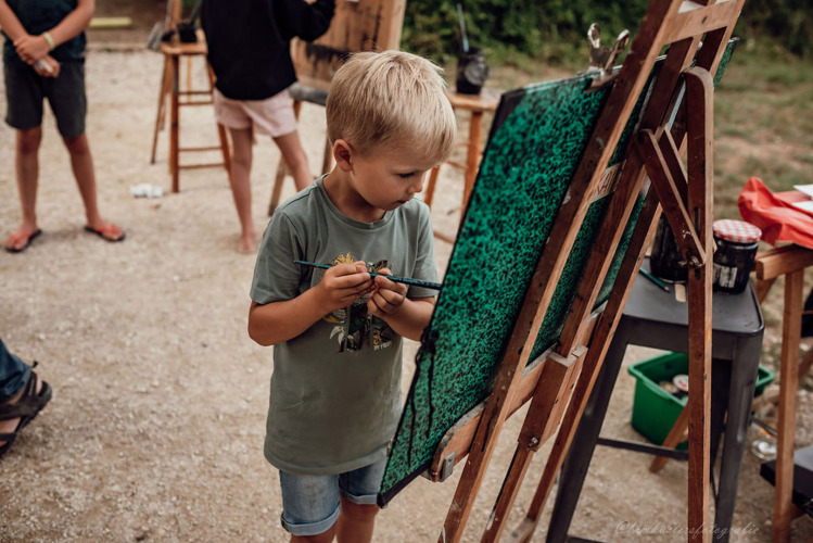 Ein kleiner Junge malt konzentriert an einer Staffelei im Freien bei Medrose Camping in Frankreich.