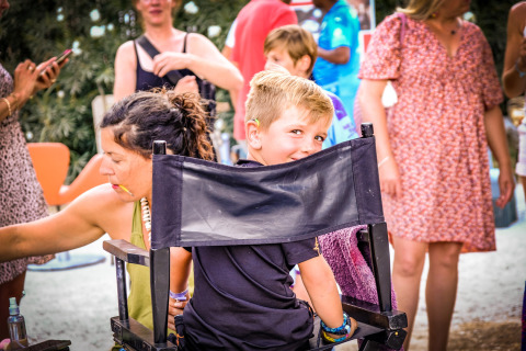 Een jongen lacht terwijl hij op een stoel zit in een levendige buitenscène op Medrose Camping, Frankrijk.