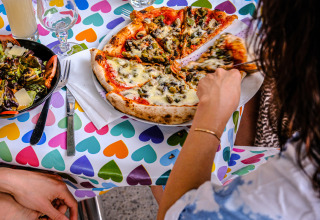 Jemand nimmt ein Stück Pizza an einem Tisch mit buntem Herzmuster bei Medrose Camping in Frankreich.
