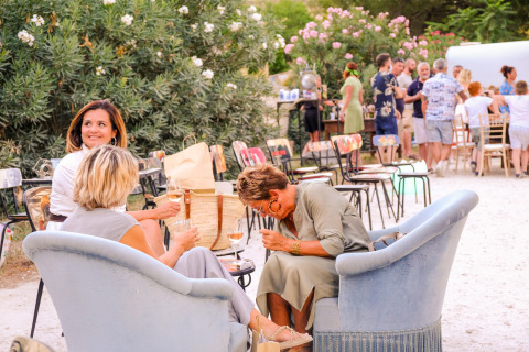 Three women relax and laugh on armchairs outdoors at Medrose Camping, with people gathering in the background.