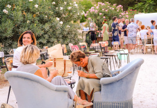 Three women relax and laugh on armchairs outdoors at Medrose Camping, with people gathering in the background.