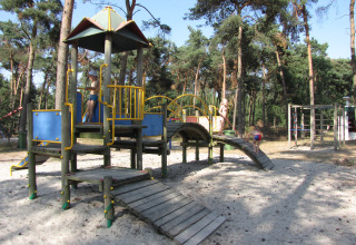Børn leger på en legeplads med træstruktur og sand, omgivet af høje fyrretræer i en skov.