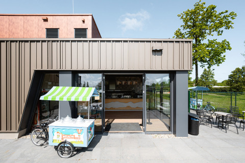 Entrada de una cafetería con carrito de helados, arquitectura moderna y ambiente soleado en RCN de Schotsman.