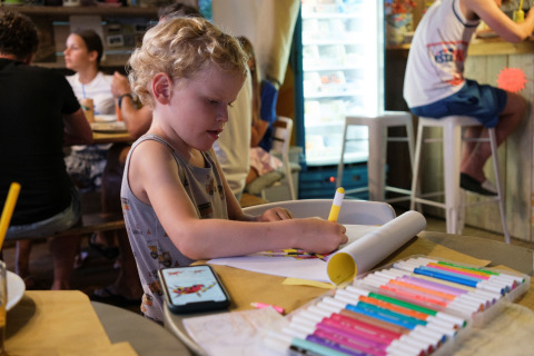 Un bambino disegna con pennarelli colorati su un tavolo in un bar con adulti sullo sfondo nelle Marche, Italia.
