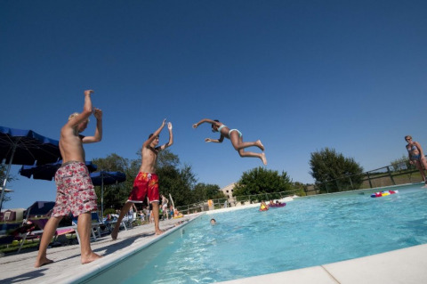 Børn leger og hopper i poolen under en klar blå himmel på Tenuta Tredici Ulivi feriepark i Marche, Italien.