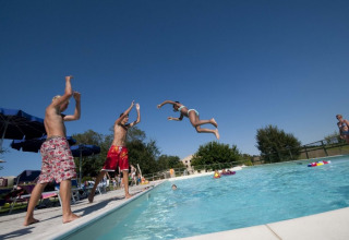Børn leger og hopper i poolen under en klar blå himmel på Tenuta Tredici Ulivi feriepark i Marche, Italien.