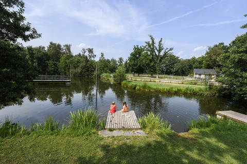 Two children sit on a wooden dock by a pond at Recreatiepark de Lucht, surrounded by lush greenery.