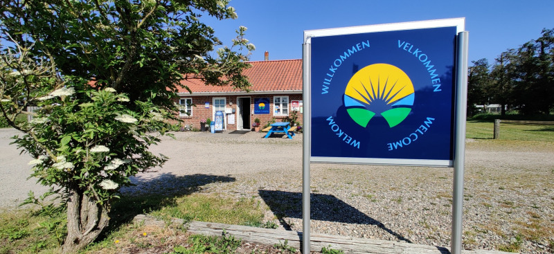 Panneau de bienvenue au Ballum Camping dans le sud du Danemark, avec bâtiment et arbre en fleurs.